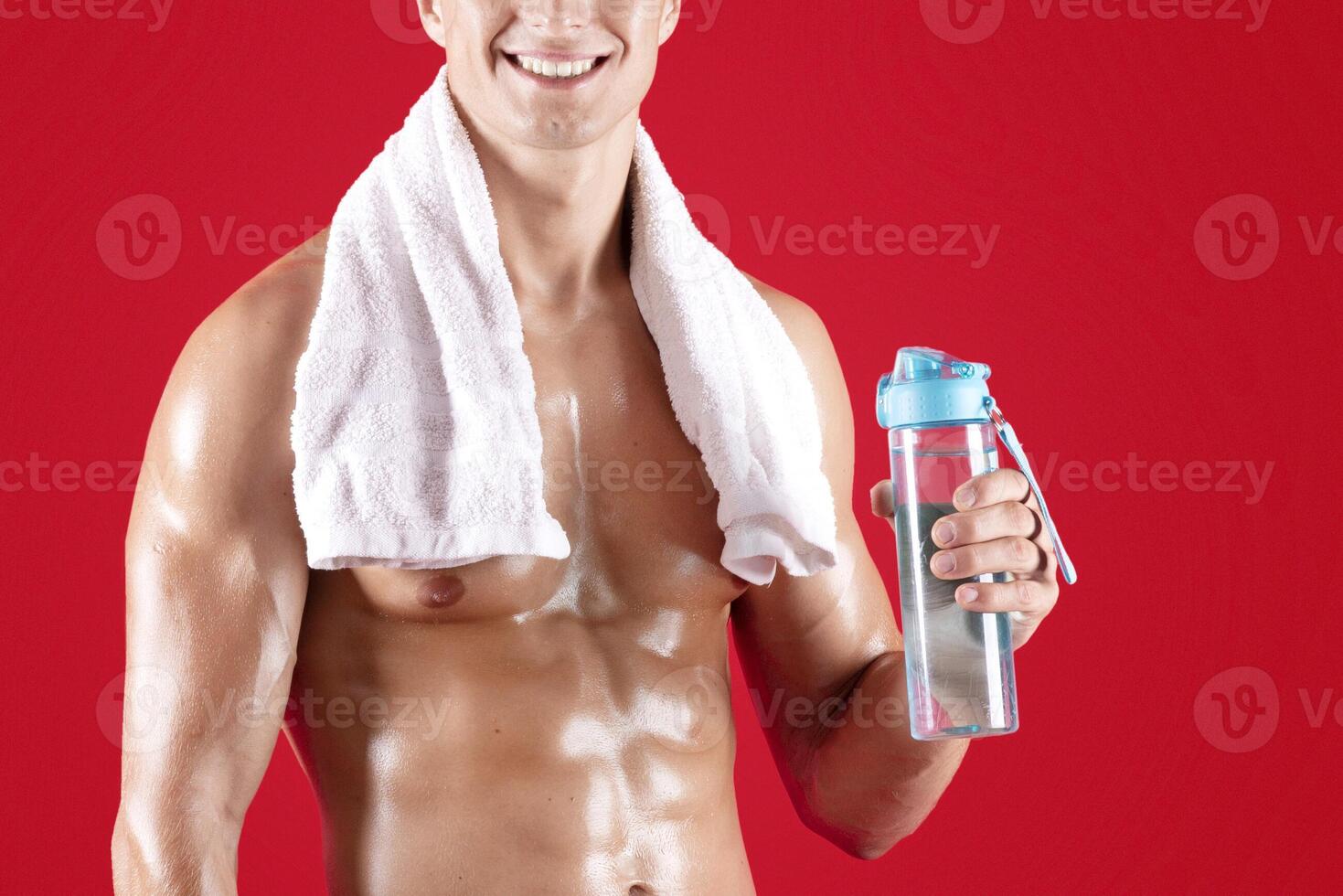 Stay hydrated during workout. Happy shirtless sportsman with towel and bottle of water on red studio background. Handsome millennial bodybuilder with naked chest looking at camera and smiling photo