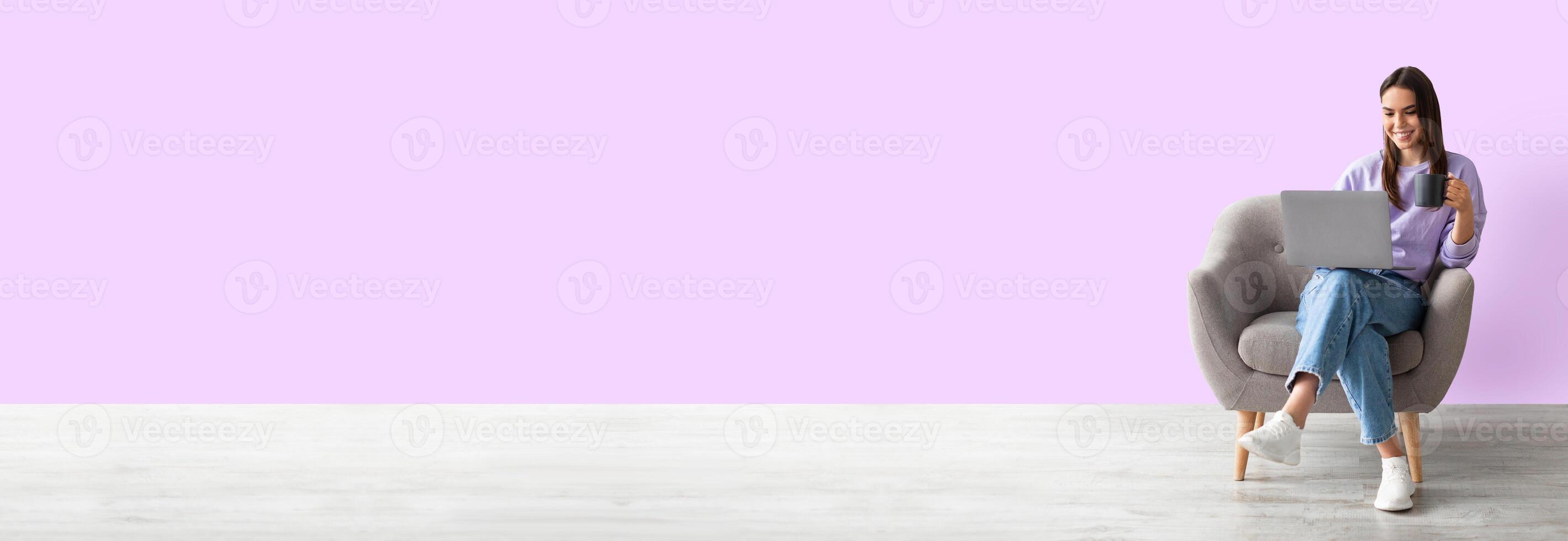 Positive young caucasian woman working online, sitting in armchair with coffee and using laptop against pink studio background, full length, panorama with copy space, mockup photo