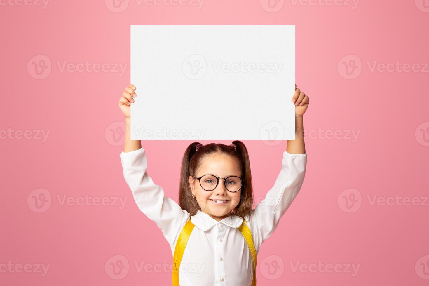 Preparation for school. Smiling little girl with backpack in glasses, holding sign with empty space, isolated on pink background, studio shoot photo