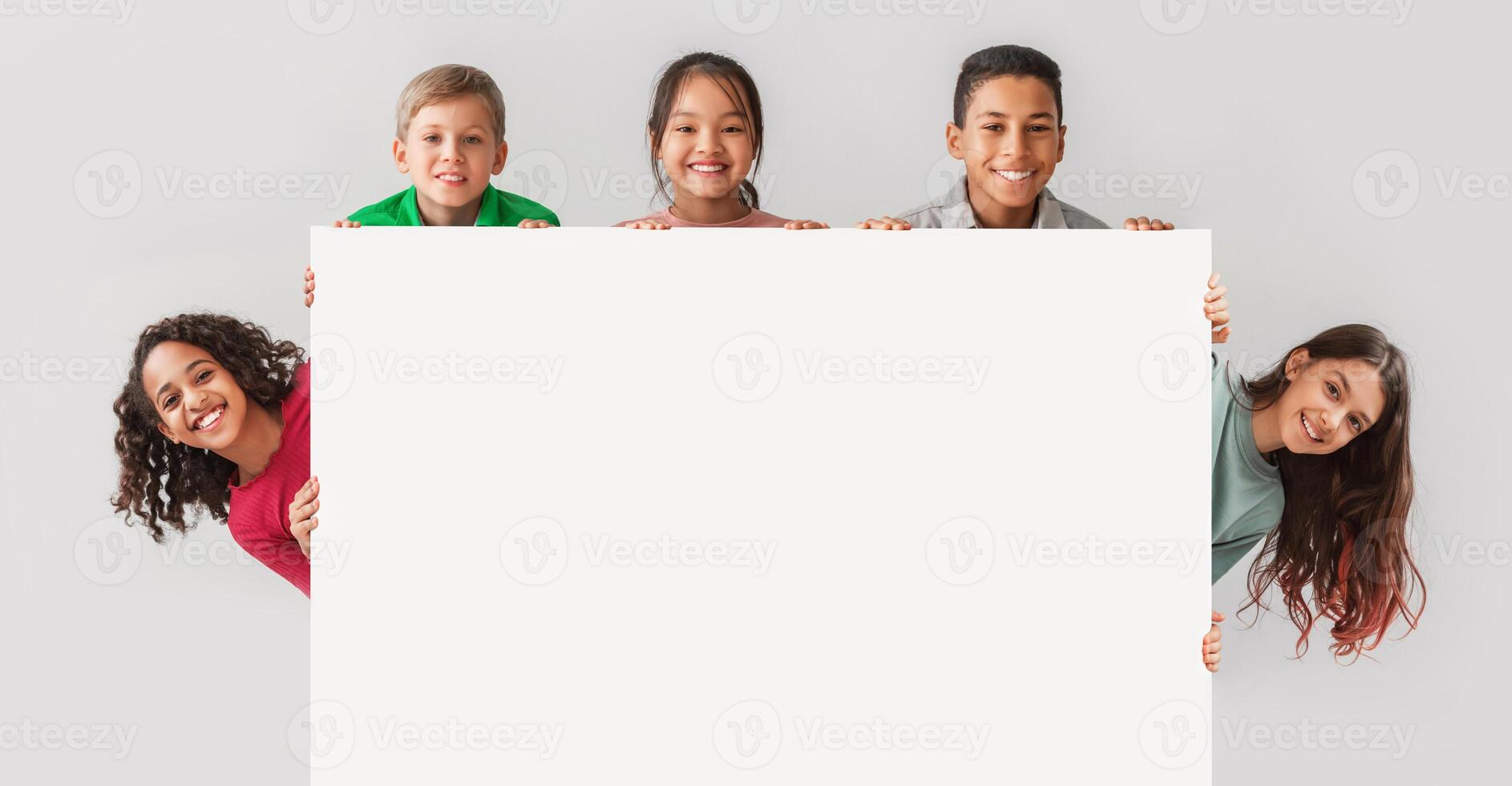 Happy Diverse Kids Hiding Behind Empty White Board Smiling To Camera Posing Over Gray Studio Background. Mixed Boys And Girls Advertising Your Text On Blank Poster. Panorama, Mockup photo