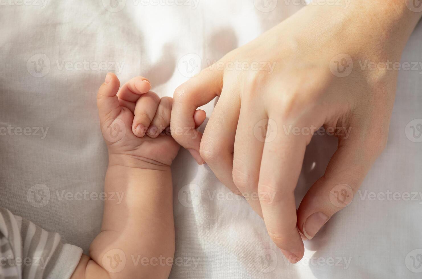 Mother Baby Connection. Newborn child holding his mom's hand while they lying in bed, touching mum's arm with his tiny fingers, top view, cropped image, closeup photo