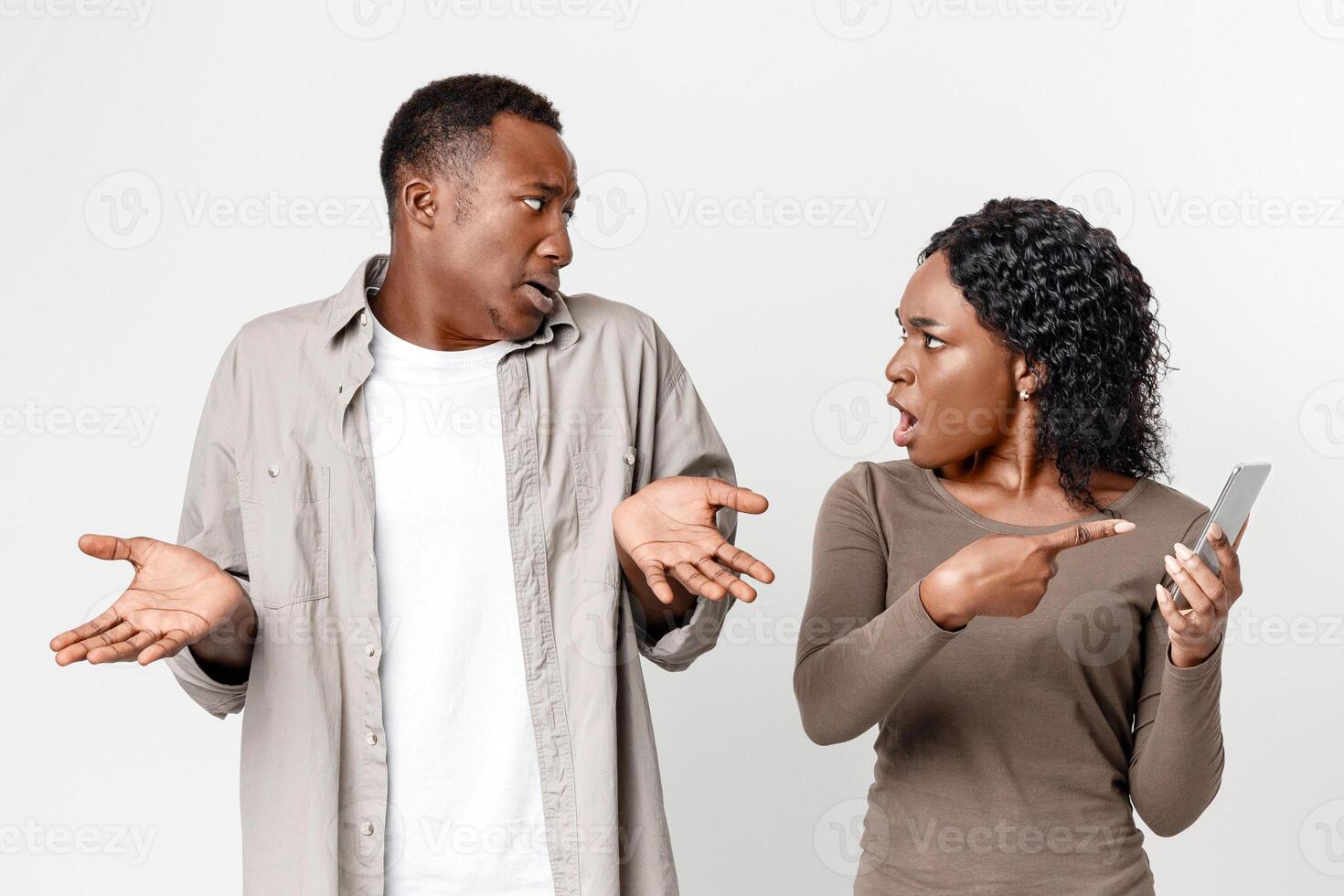 Gadget and social media addiction, cheating in relationships. Black woman checking her boyfriend mobile phone, showing provocative messages and yelling at man, gray studio background photo