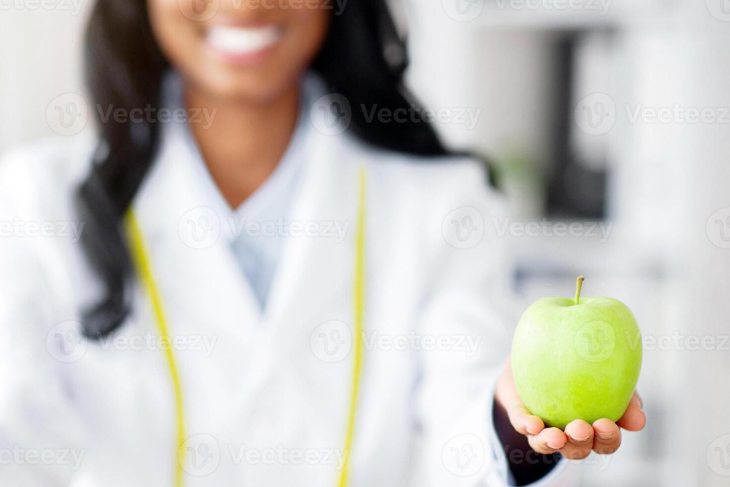 Closeup view of dietitian's hands holding fresh apple and donut at table in medical office. Cropped view of black nutritionist offering choice between healthy and junk foods. Dieting concept photo
