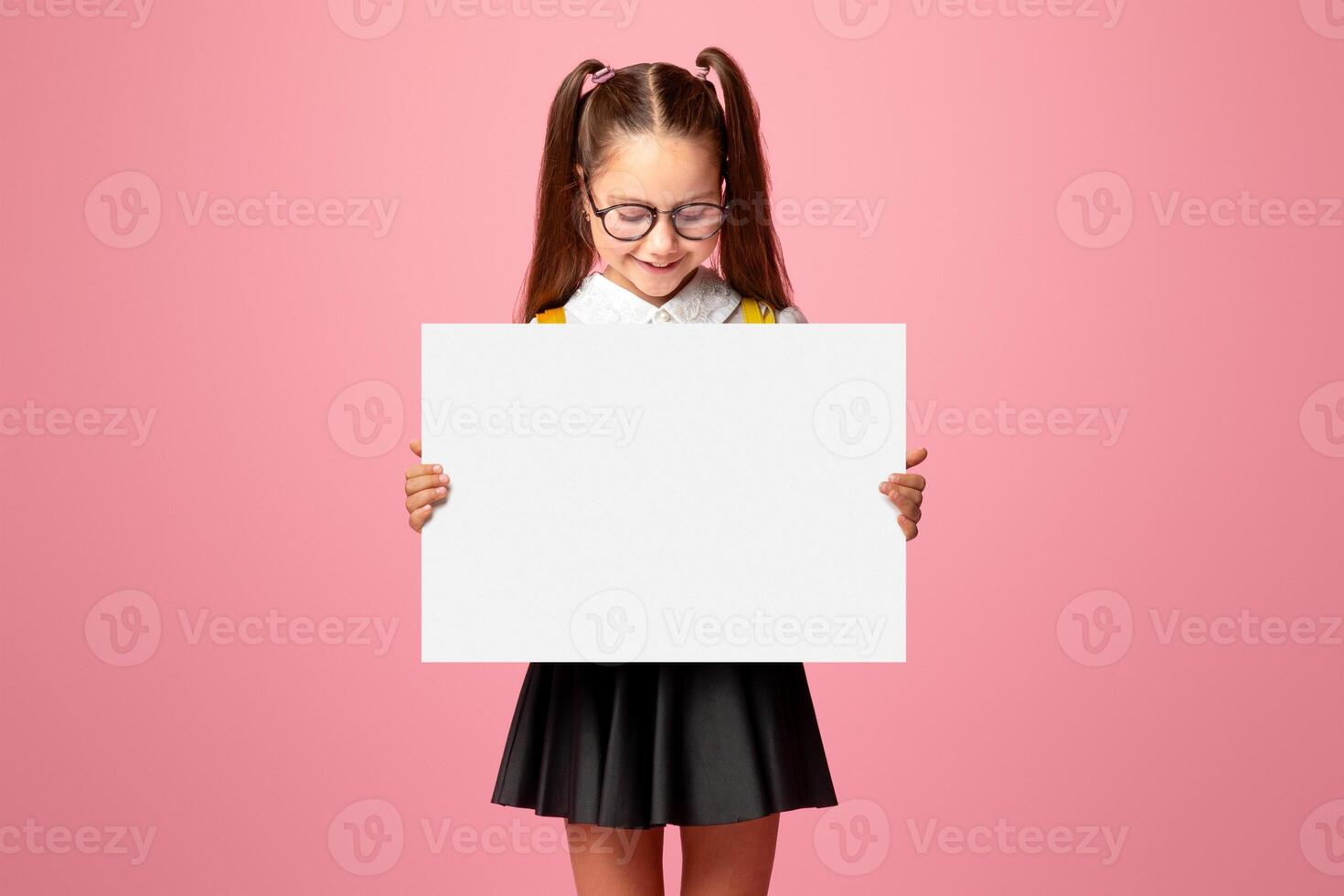Private schooling. Small child girl in glasses and school uniform holds table with empty space and looks at it, isolated on pink background photo