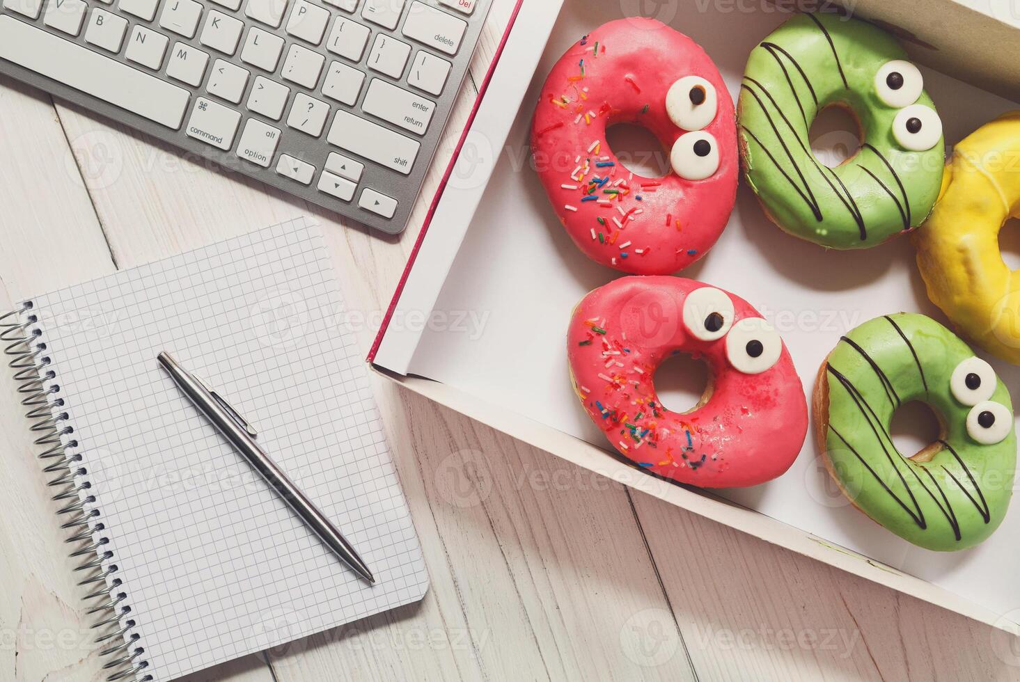 Box of colorful glazed donuts with funny eyes on the office desk near computer keyboard and notepad with pen. White rustic wooden table with business lunch snack dessert top view photo
