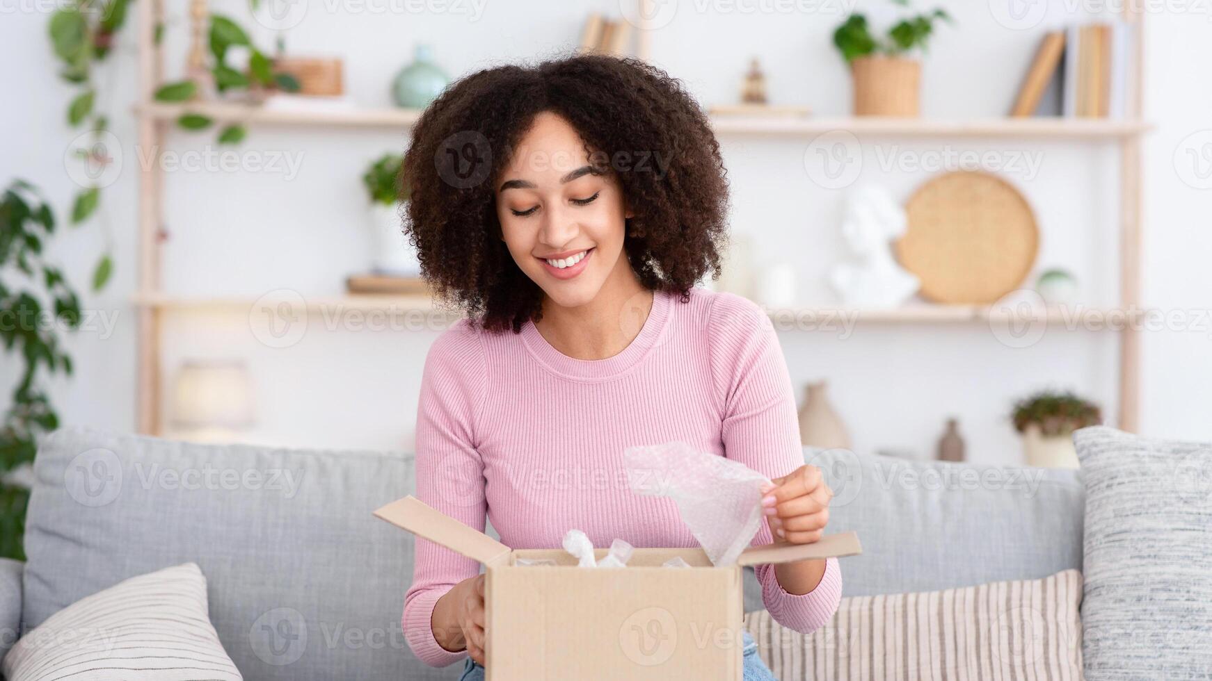 Online shopping, pleasure of buying, parcel delivery, good service. Happy interested young african american lady opens cardboard box and looks inside, sits on sofa in living room interior, free space photo