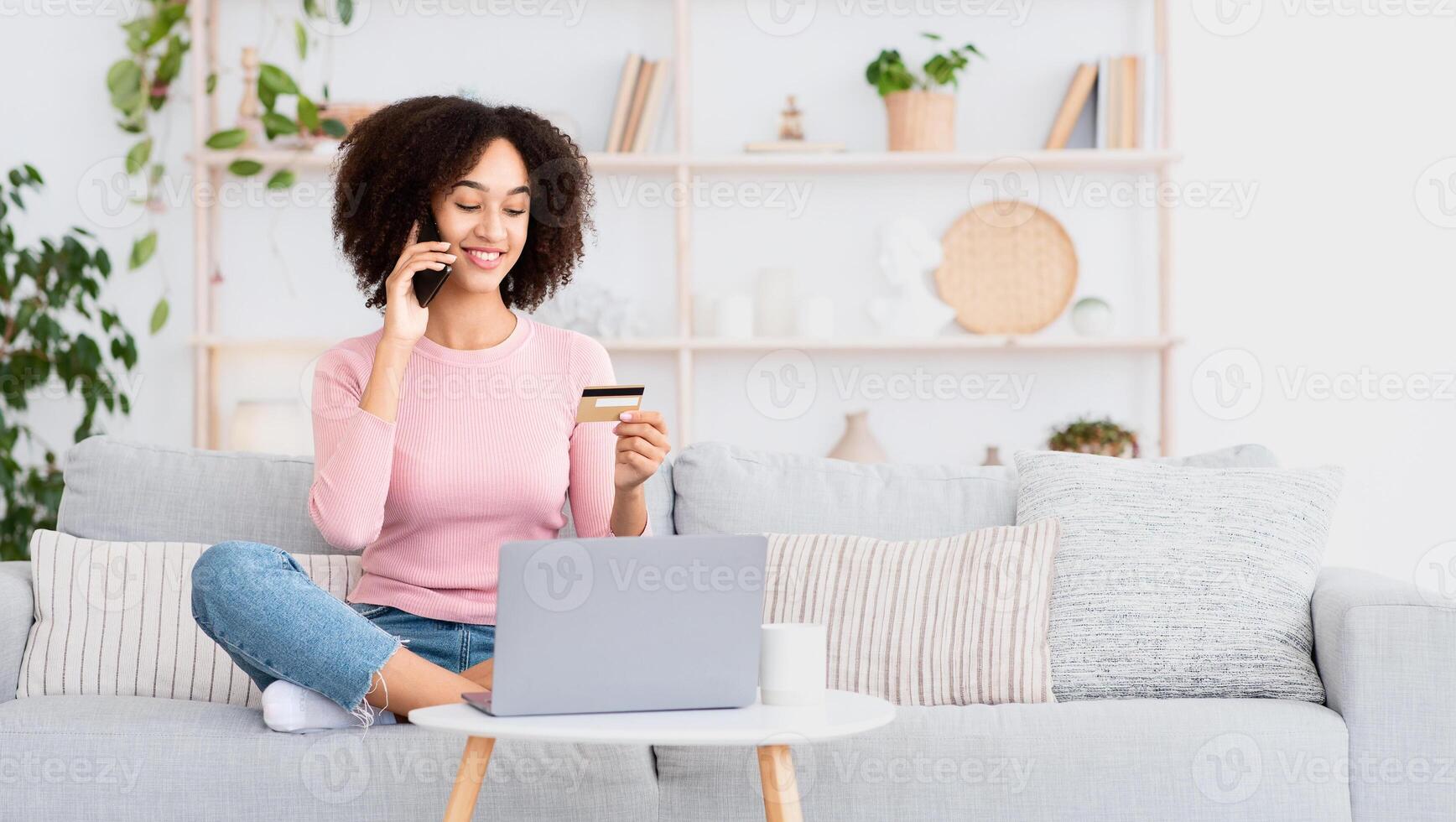 Online service, customer support for online shopping and purchase payment. Cheerful millennial african american woman sits on couch with laptop, calling on phone and holds credit card in living room photo