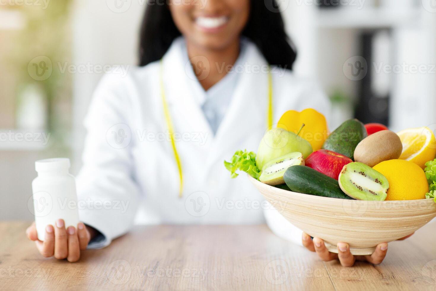 Happy black dietitian offering choice between fresh products and tablets at clinic. Young nutritionist holding bowl of fruits with veggies and jar of pills or vitamin supplements. Healthy eating photo
