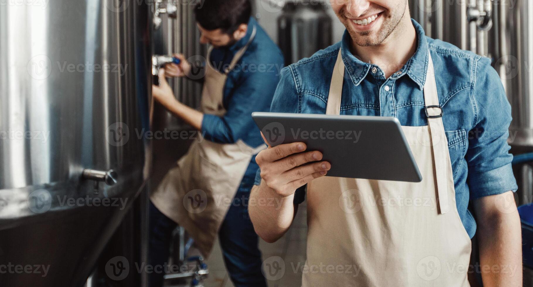 Beer production process at brewery. Man operating brewing equipment at beer making factory and using digital tablet. Cheerful young handsome men in aprons check data on large kettle with craft drink photo