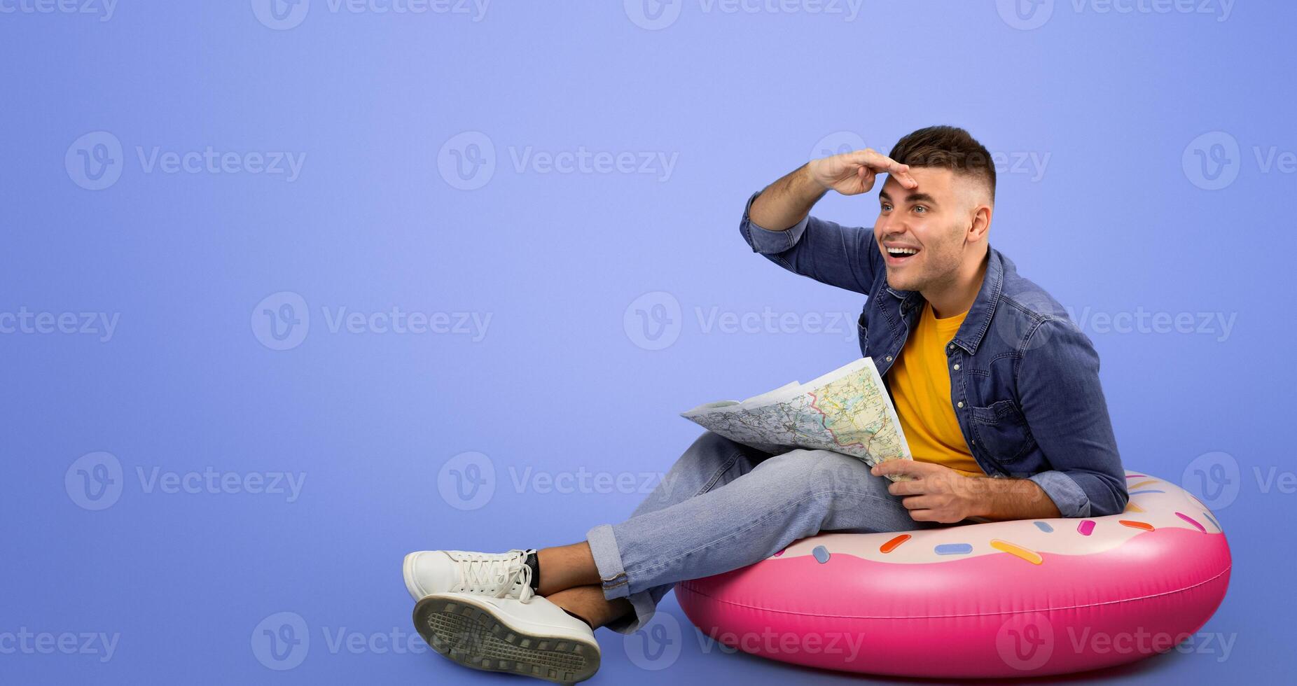 Funky young guy in casual wear sitting on inflatable ring with city map and looking forward, blue studio background. Full length portrait of traveler dreaming about his summer vacation photo