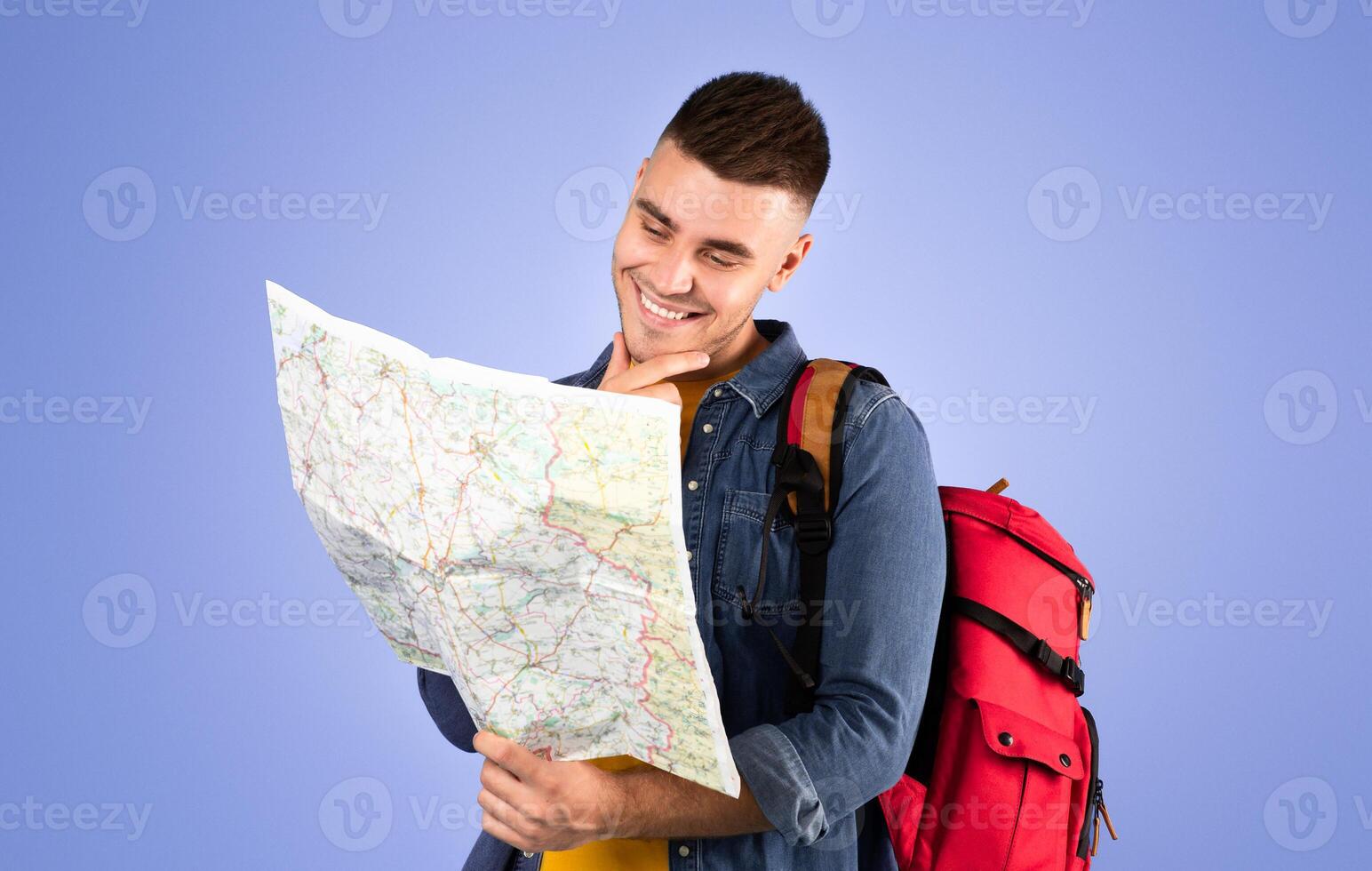 Portrait of happy young traveler with backpack and map on blue background. Positive millennial hiker planning his weekend camping trip route photo