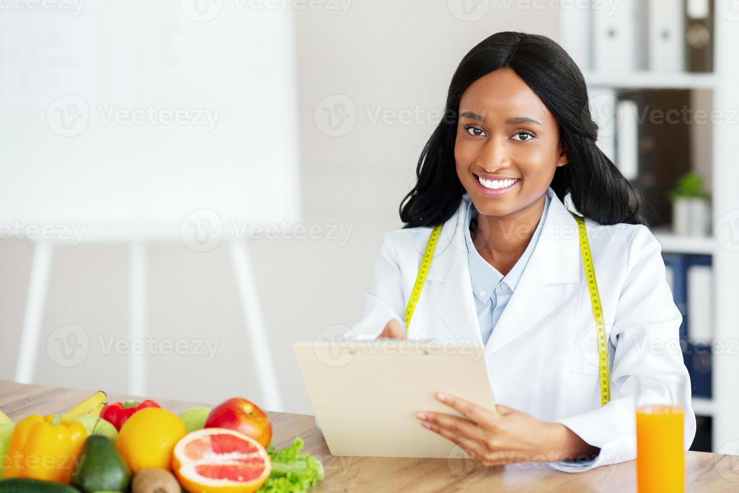 Millennial black nutritionist with clipboard creating plant based meal plan for patient at weight loss clinic, free space. Cheerful female dietitian prescribing healthy diet to client at office photo
