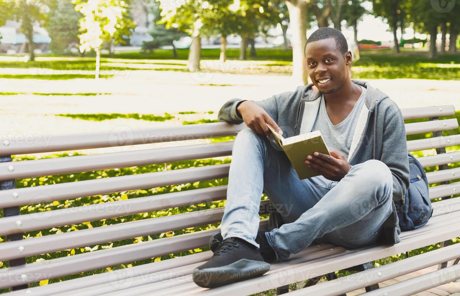 Smiling african-american student reading a book in the park, studying and preparing for exams at the university or college. Technology, communication, education and remote working concept, copy space photo