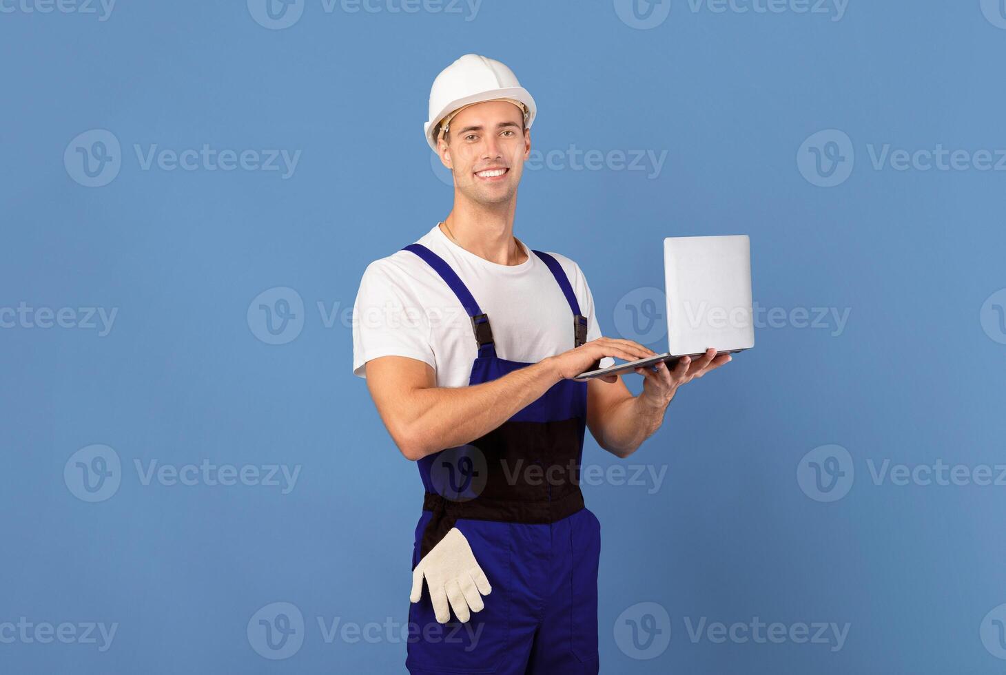 Smiling young man in coveralls and hard hat using laptop computer on blue studio background, handsome male worker ordering building instruments and materials for flat renovation online, copy space photo
