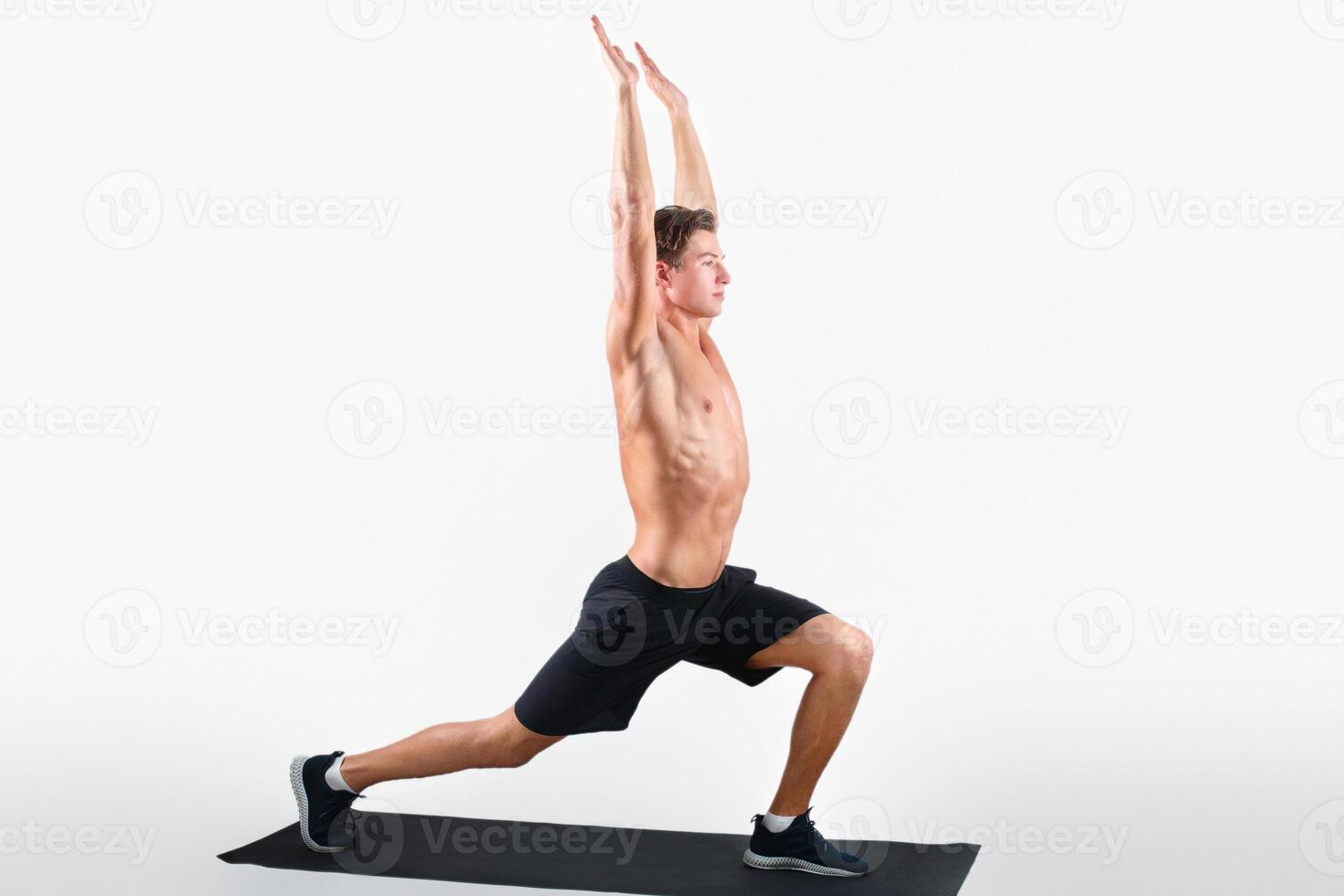 Healthy lifestyle and wellbeing concept. Flexible young guy doing yoga or pilates exercises on light studio background. Peaceful millennial man standing in warrior asana on mat photo
