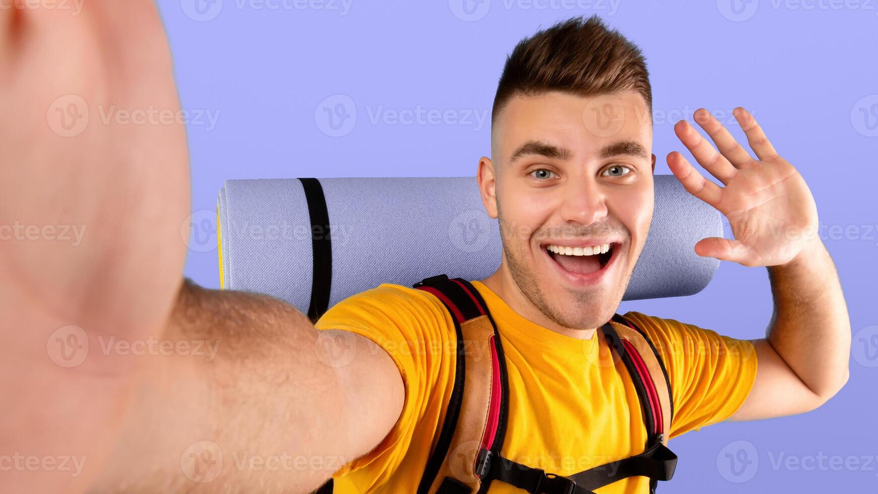 Funky millennial guy with camping gears taking selfie and waving to smartphone camera over blue studio background. Cheerful young hiker taking cool photo of himself before tourist trip