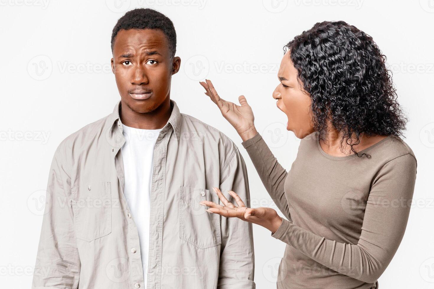 Furious african american young woman yelling at her innocent and upset boyfriend. Angry black lady suspecting her husband in cheating, grey studio background. Distrust, cheating in relationships photo