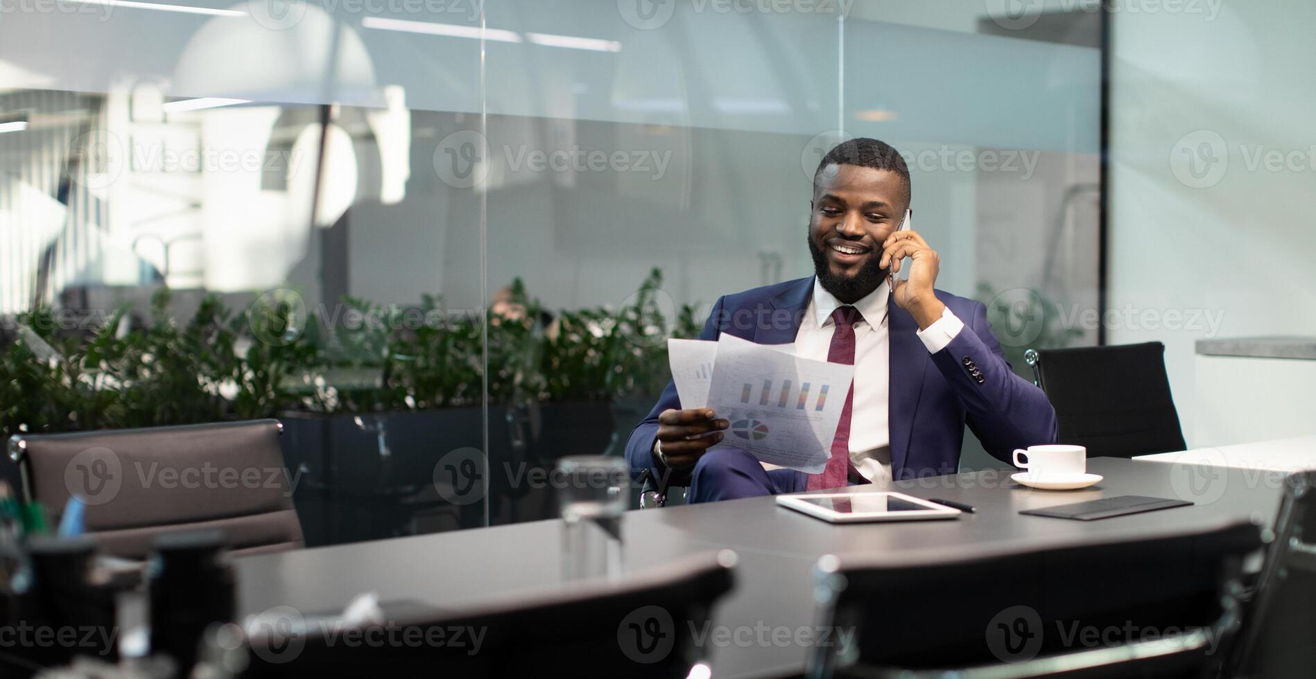 Wealthy black guy in suit entrepreneur having phone conversation with his assistant or business partner, sitting at table with digital tablet on, looking at documents, panorama with copy space photo