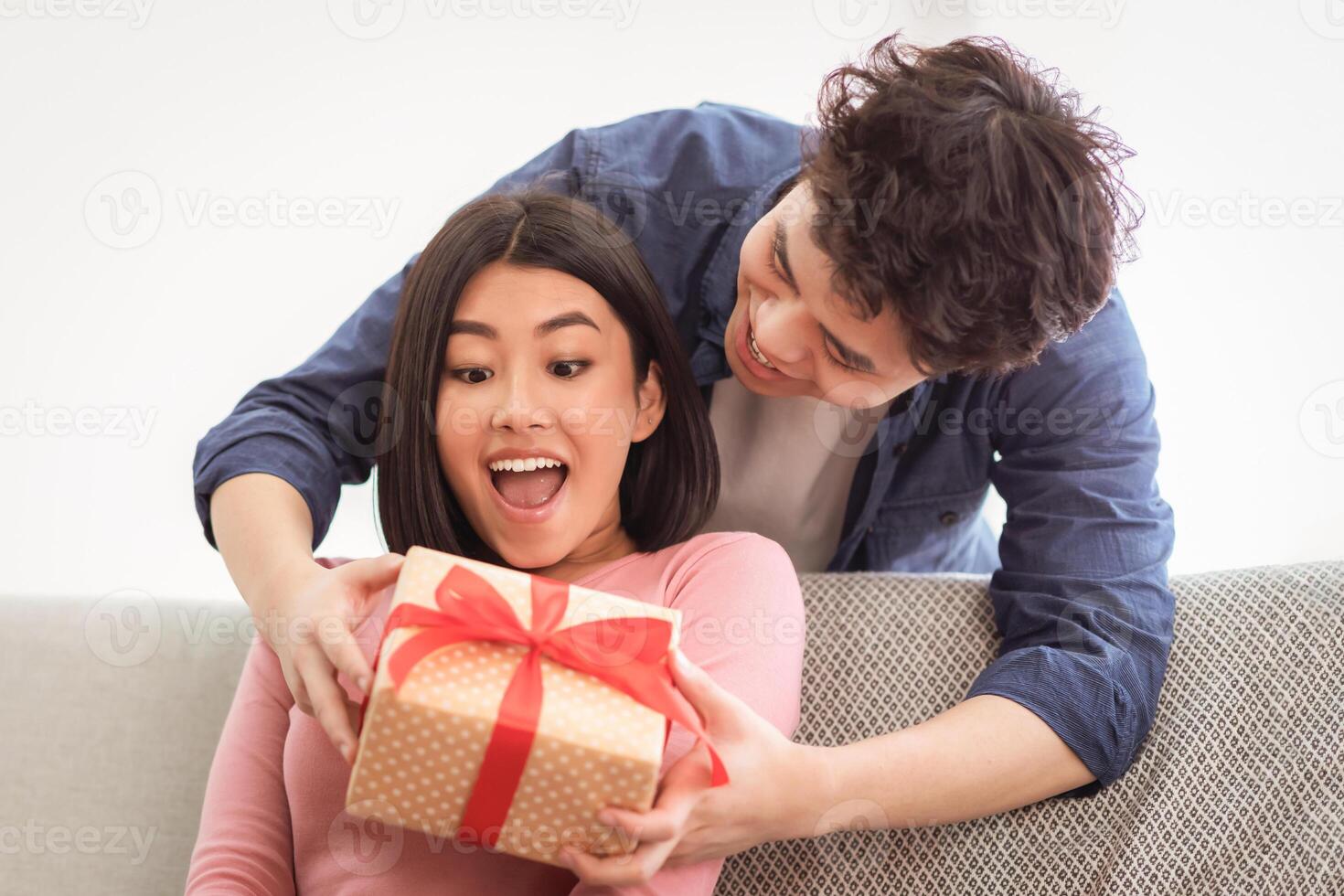 Asian Husband Giving Wrapped Christmas Gift To Wife Surprising Her With Xmas Present Sitting On Sofa At Home. Family Holidays Celebration, Birthday And X-Mas Gifts And Presents. Free Space photo