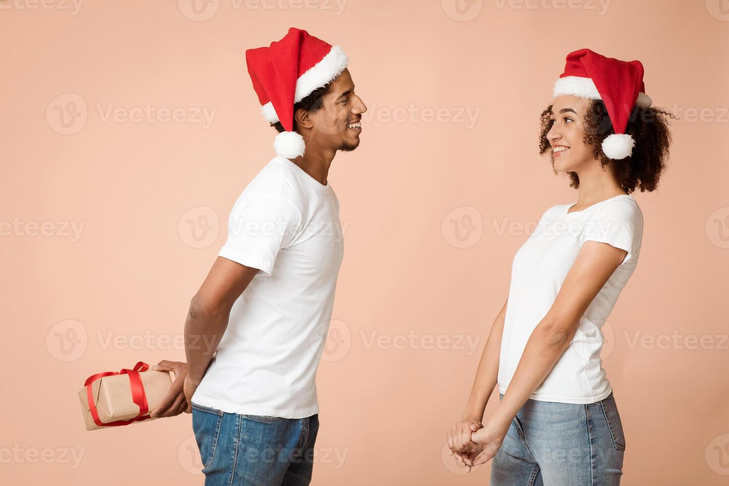Happy young couple celebrating New year and Christmas on party. African american man in hat with smiling holding box, lady in white t-shirt waiting for gift isolated on light, studio shot, free space photo