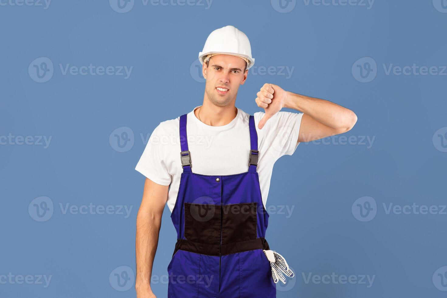 Disappointed Handyman In Hardhat And Coveralls Showing Thumb Down Gesture At Camera, Displeased By Unprofessional Repair Services, Standing Over Blue Studio Background With Copy Space photo