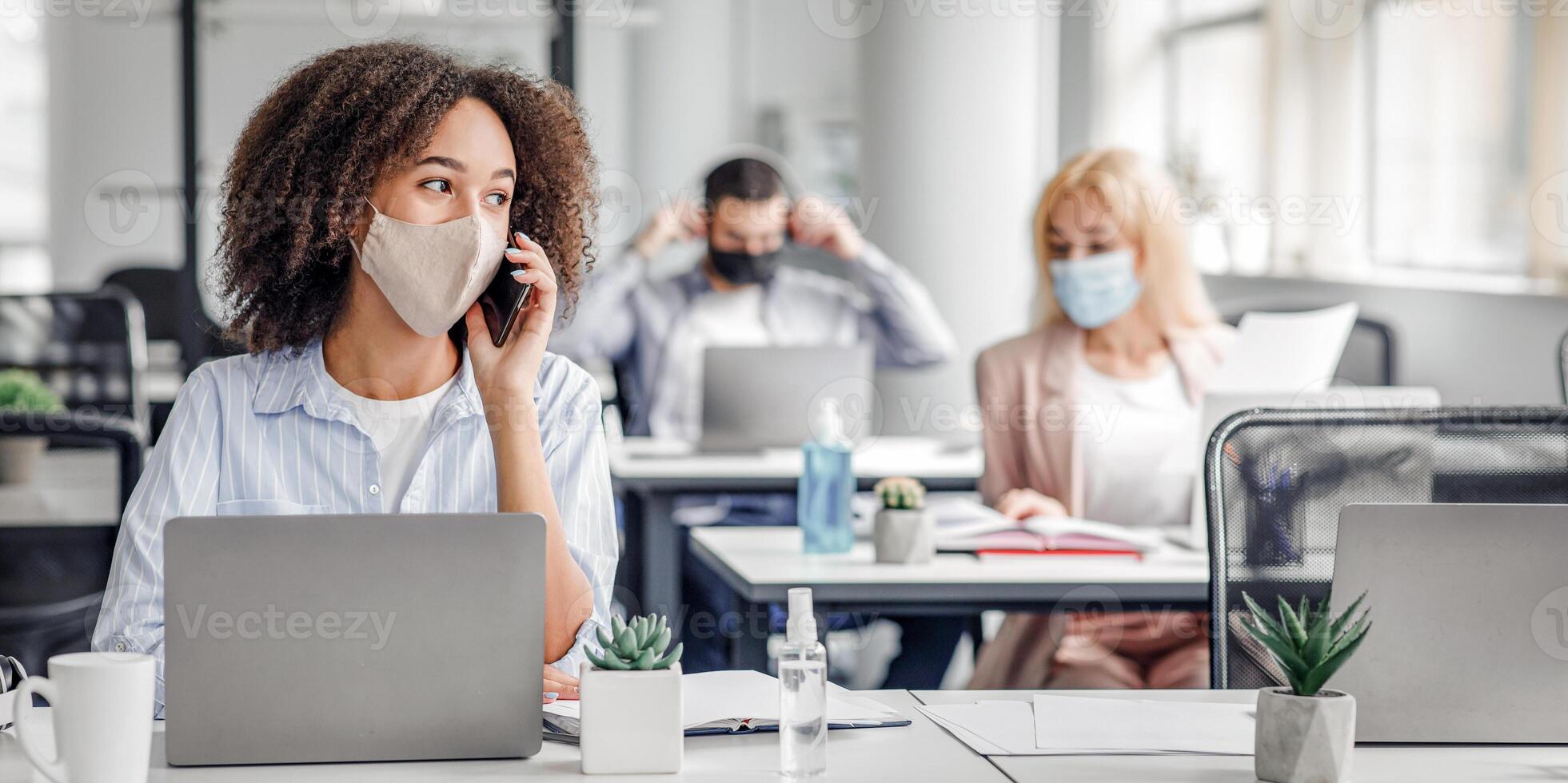 Working with clients remotely after returning from quarantine. African american woman in protective mask speaks on phone, sitting at table with laptop in modern office interior with colleagues, empty space photo
