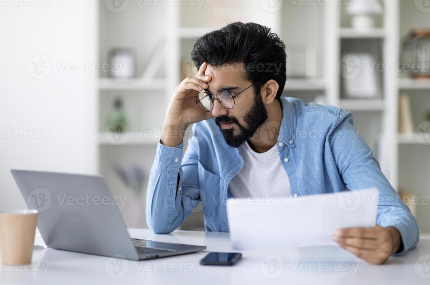 Stressed Young Indian Man Looking At Laptop Screen And Holding Documents, Focused Millennial Eastern Male Cheching Business Reports While Working Remotely At Home Office, Closeup Shot With Free Space photo