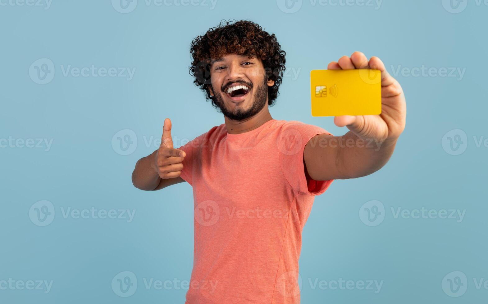 Happy curly milennial hindu guy in pink t-shirt pointing at yellow credit card in his hand and smiling, shopaholic enjoying unlimited shopping, blue studio background, copy space photo