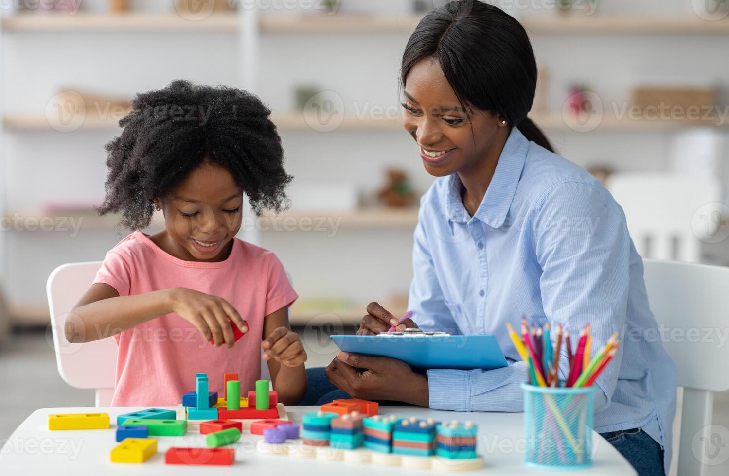 Female psychotherapist friendly pretty young black woman working with little kid at office, sitting by preschooler girl playing with bricks, taking notes and smiling. Child therapist concept photo