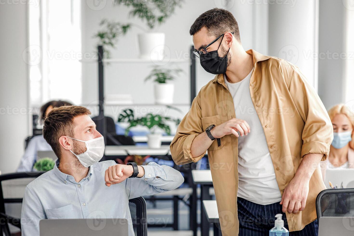 Safe hello at work during COVID-19 epidemic and social distance. Young men in protective masks greet with elbows at workplace with laptop and antiseptic in modern office interior, copy space photo
