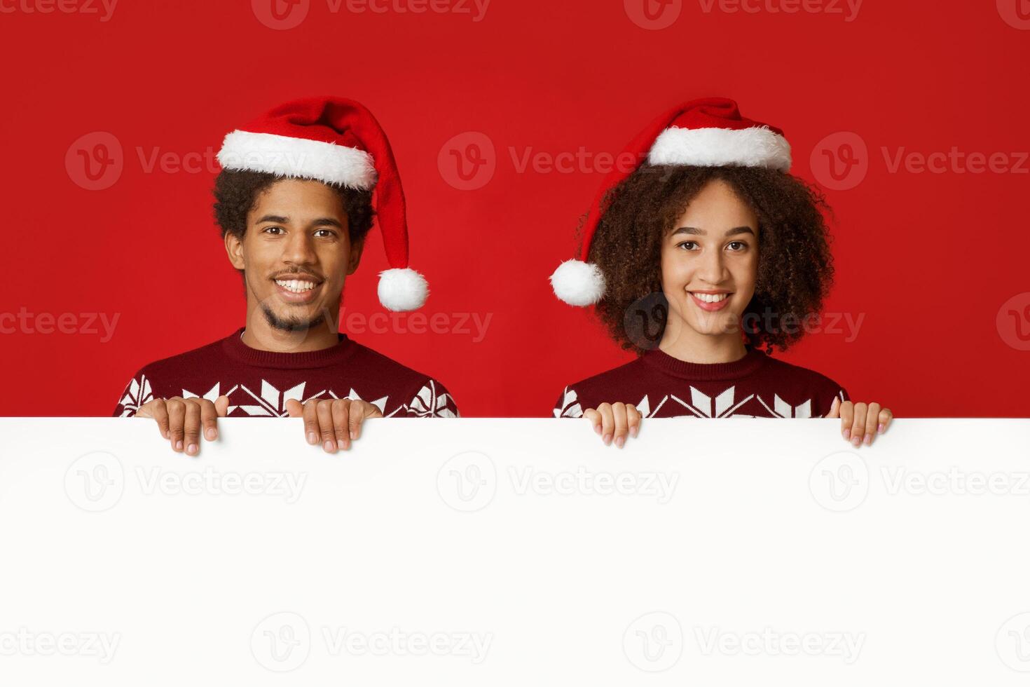 Winter mood and Christmas celebration. Cheerful young african american friends or family in Xmas sweaters and hats, holds white poster with empty space, isolated on red background, studio shot photo