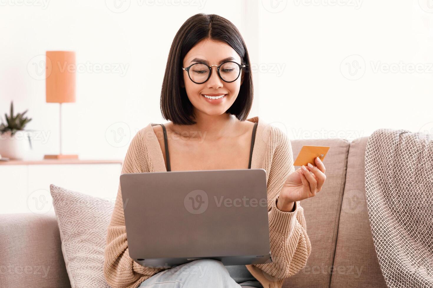 Online Banking Concept. Happy asian woman in eyeglasses making purchases on internet with laptop, holding credit card to pay. Easy Shopping. Smiling lady using computer sitting on couch, copy space photo
