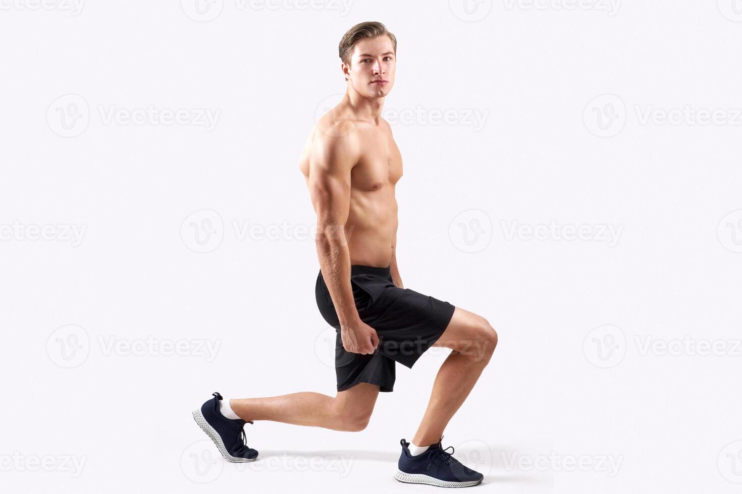 Strong young guy with sexy muscular body doing lunges, warming up before workout over light studio background. Full length portait of handsome shirtless man exercising, stretching legs photo