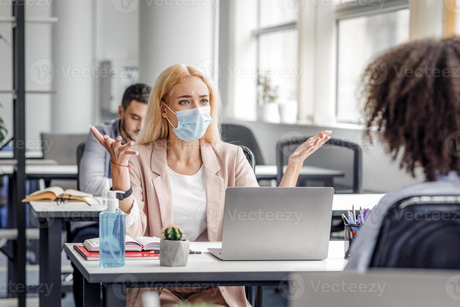 Conflict at work and problems with project. Angry woman in protective mask at workplace with laptop yelling at african american lady and gestures in interior of modern office, copy space photo