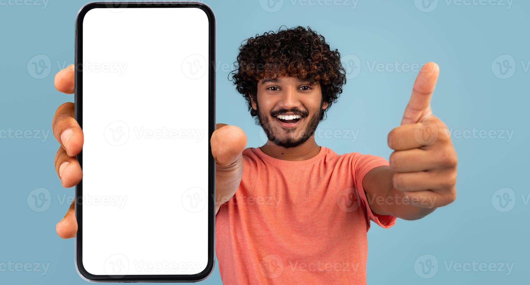 Handsome indian millennial guy showing brand new smartphone with empty white screen and thumb up, smiling at camera over blue studio background, mockup, mobile application concept, panorama photo