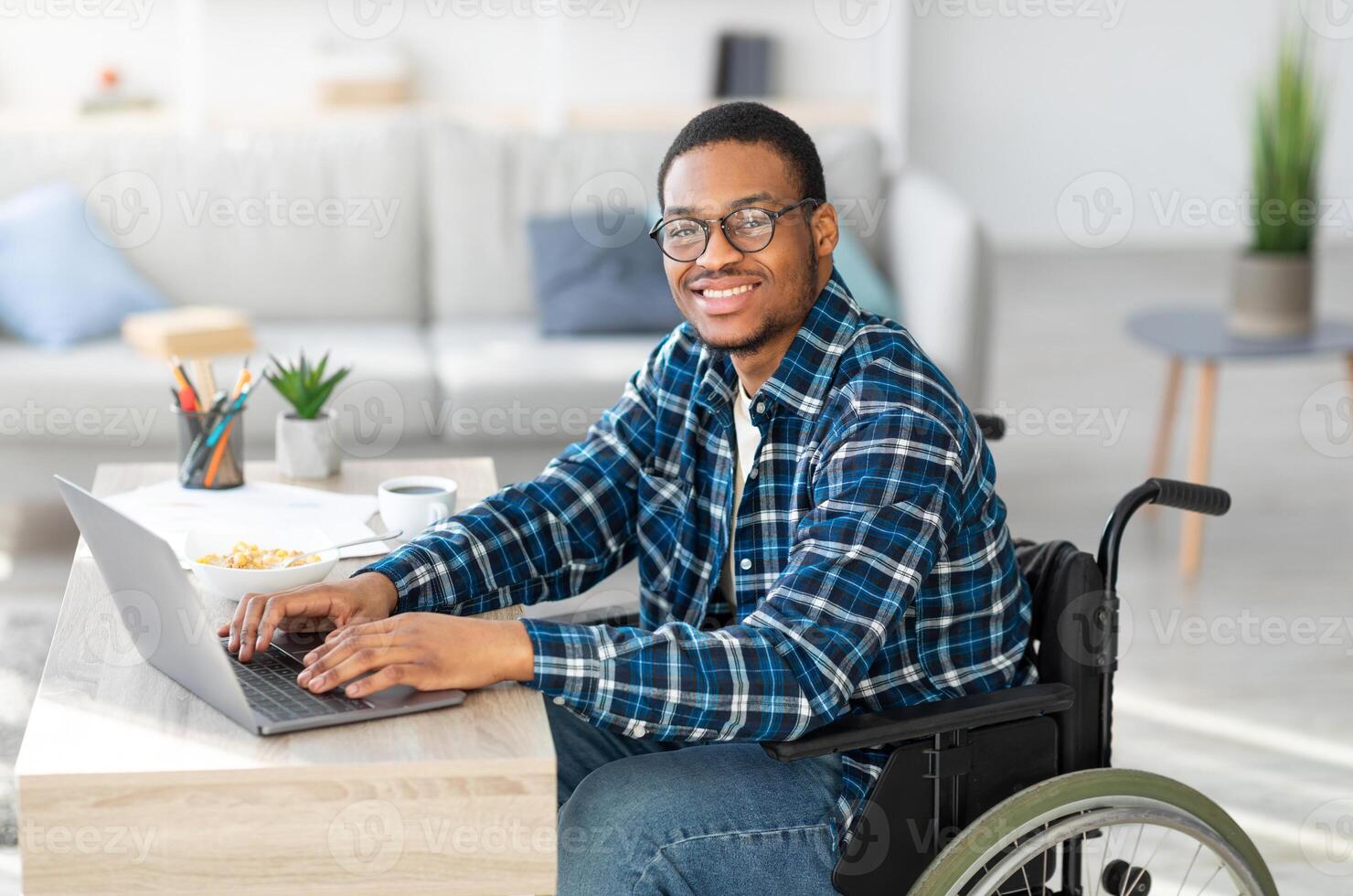 Cheerful handicapped black man in wheelchair using laptop for online job from home, having business meeting on web. African American disabled freelancer working on remote project photo