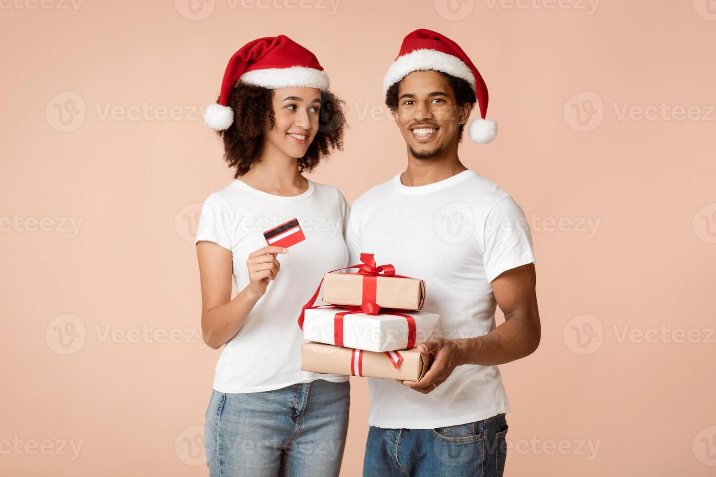 Easy and quick gifts for Christmas and New Year with online shopping. Cheerful young african american couple in Xmas hats holds many gifts and credit card, isolated on light background, studio shot photo