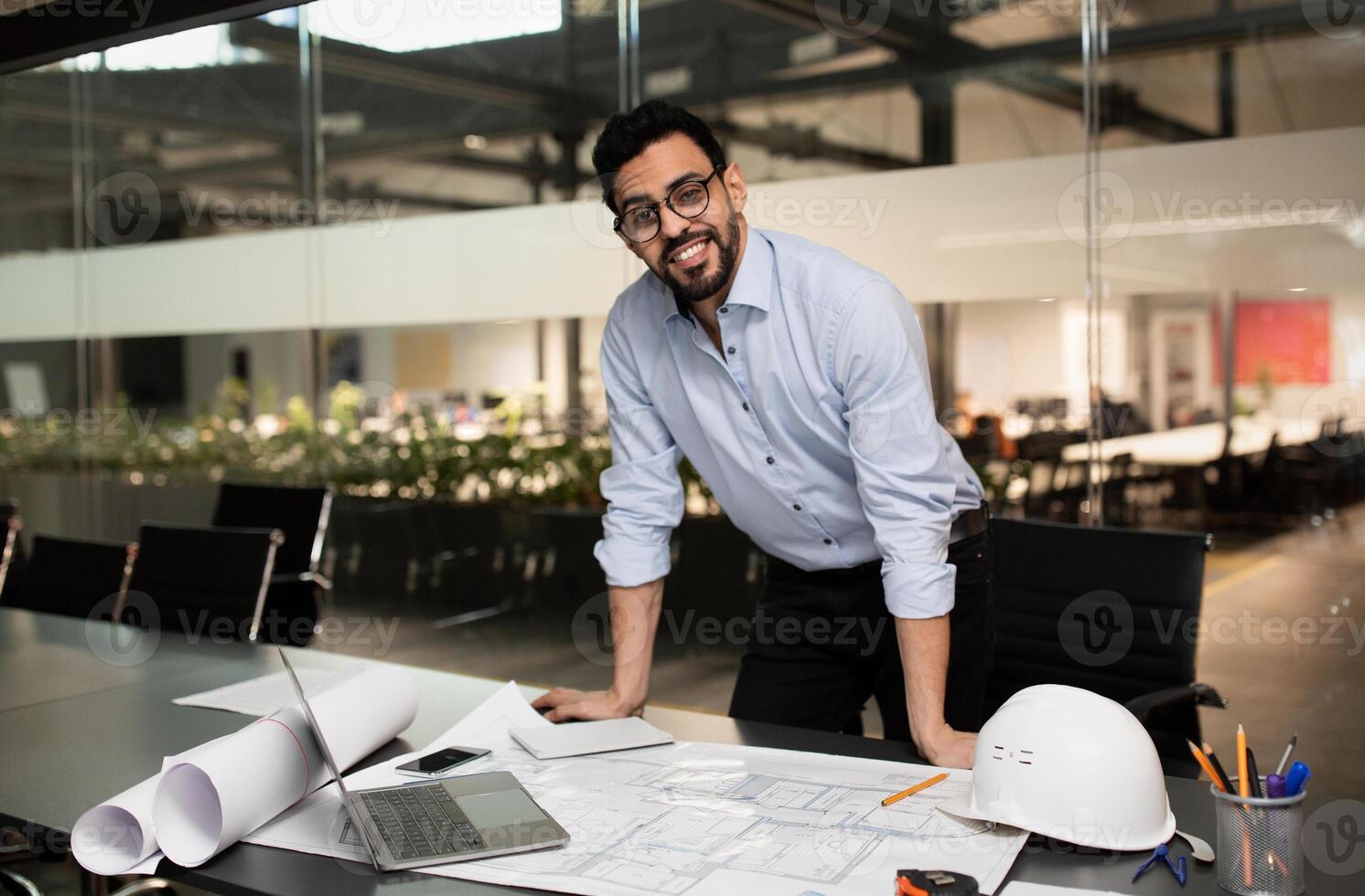 Smiling millennial attractive muslim engineer builder in glasses working on desk with project drawings in office factory interior, free space. Designer and professional, production analysis at plant photo