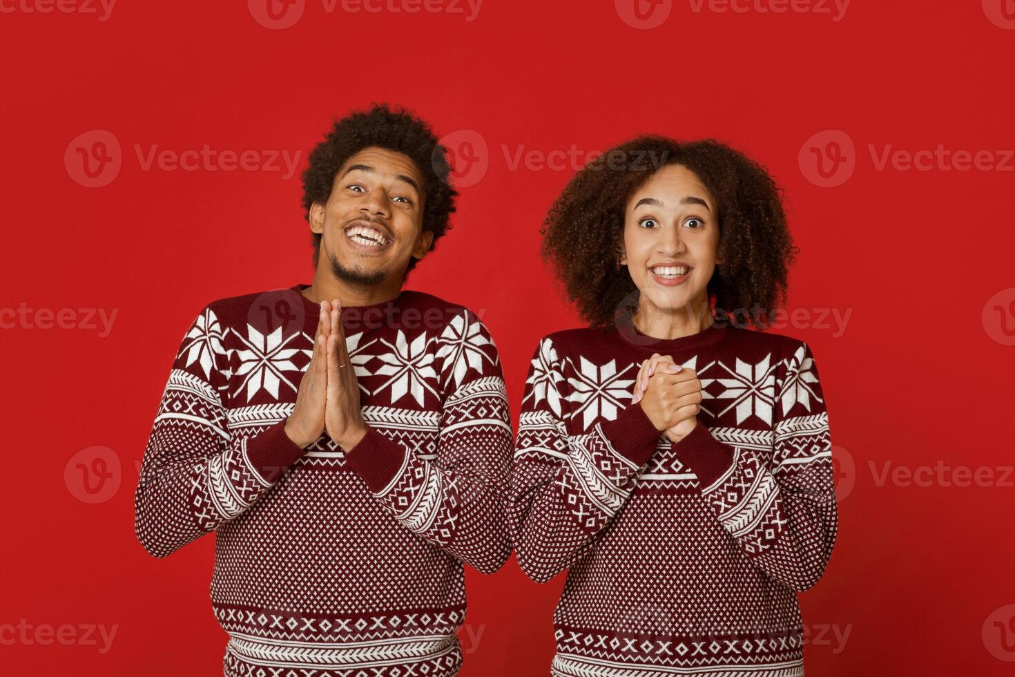 Good luck and great wish for New Year and Christmas. Excited african american couple in sweaters, smiling and hoping for desire, folded hands in prayer, isolated on red background, studio shot photo