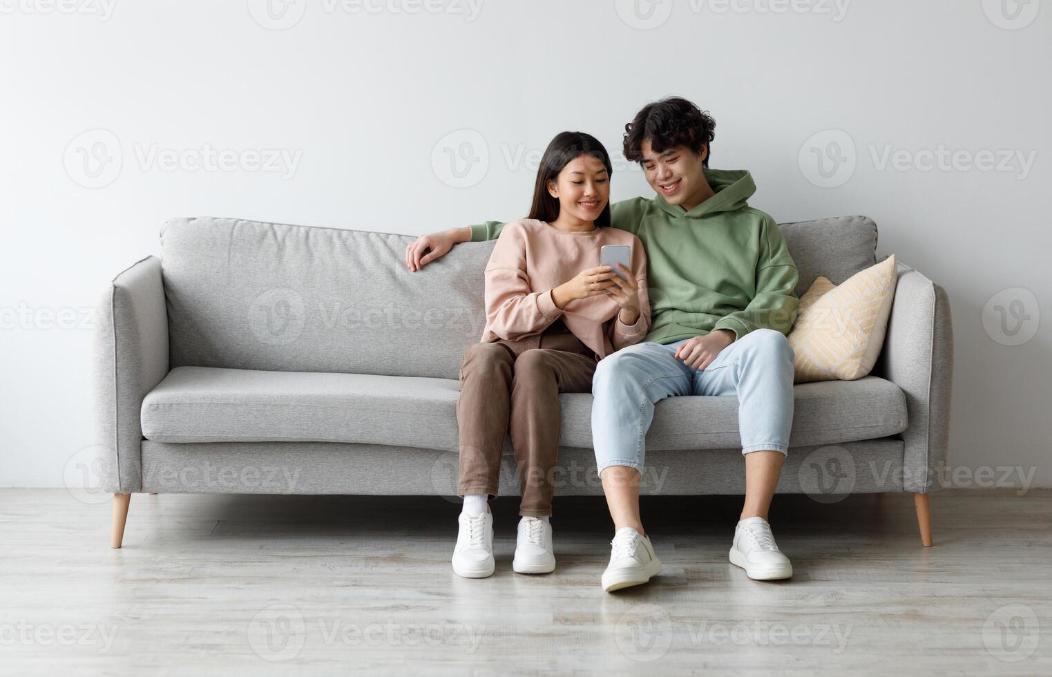 Happy young Asian couple using cellphone, browsing internet, chatting online, relaxing on sofa at home, free space. Cheery millennial family checking new mobile application, resting on couch together photo