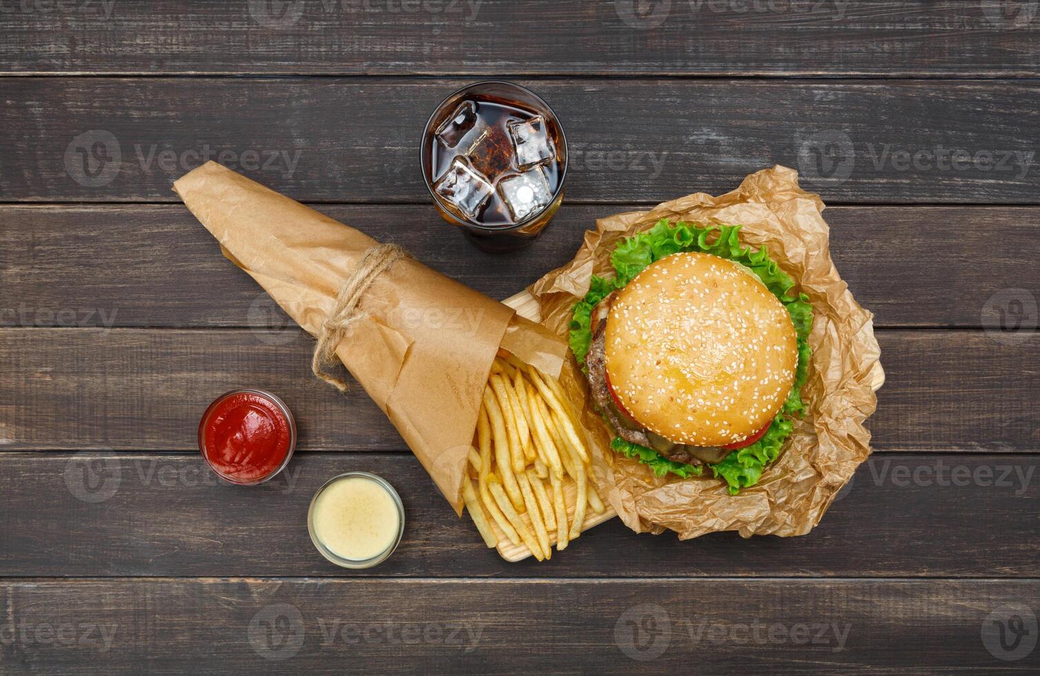 Fast food dish top view. Meat burger, potato chips and glass of cola drink with ice on wood. Takeaway composition. Wrapped French fries, hamburger, mayonnaise and ketchup sauces on wooden desk. photo
