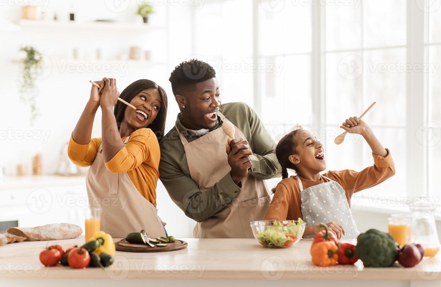 Positive Black Family Of Three Cooking And Having Fun Singing Favorite Song Holding Spoons Preparing Dinner Together Standing In Modern Kitchen Indoor. Weekend Leisure, Healthy Food Recipes Concept photo