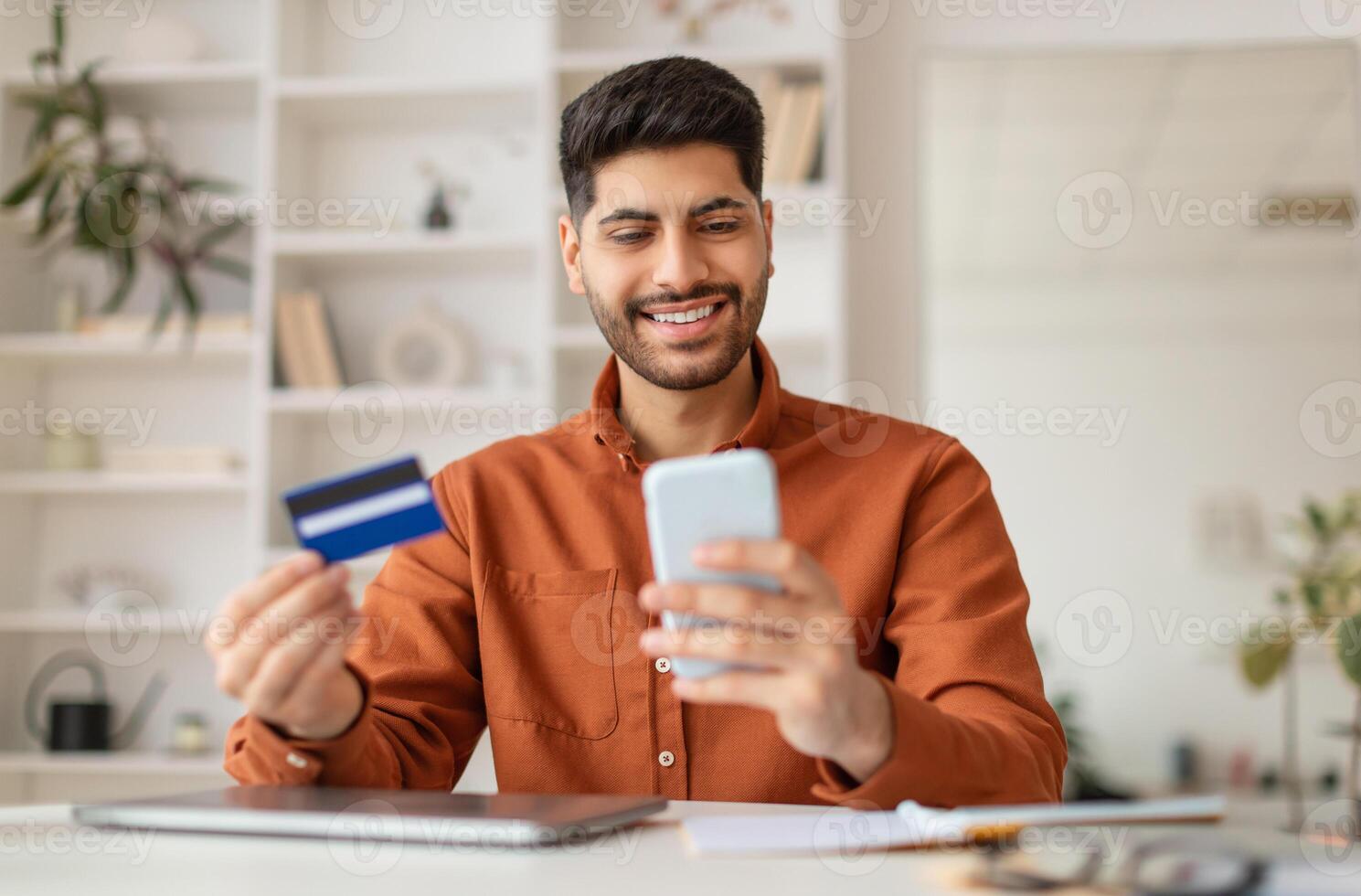 Internet Mobile Shopping Concept. Positive Middle Eastern man using cell phone looking at screen holding credit card purchasing things online sitting at desk at home office, buying gifts in web store photo