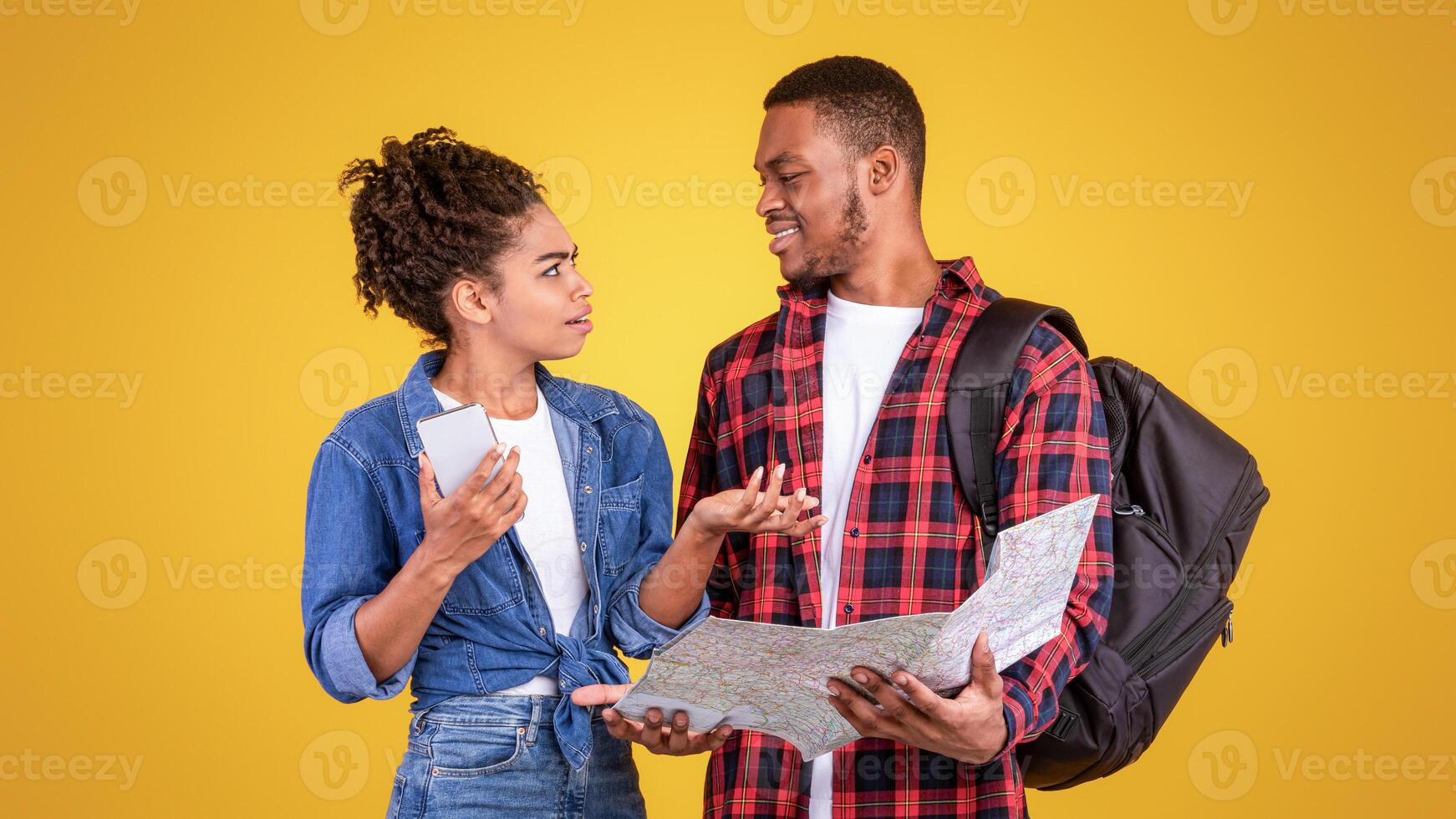 Black Couple Having Quarrel Planning Vacation Holding Smartphone And Tourist Map Choosing Journey Destination Standing On Yellow Studio Background. Travel Navigation Problems Concept photo
