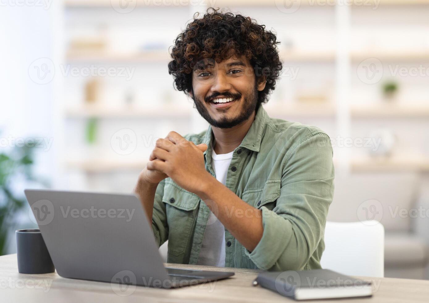 Cheerful attractive indian guy in casual outfit programmer posing at workdesk at office, eastern freelancer sitting in front of computer at home, working online, smiling at camera, copy space photo