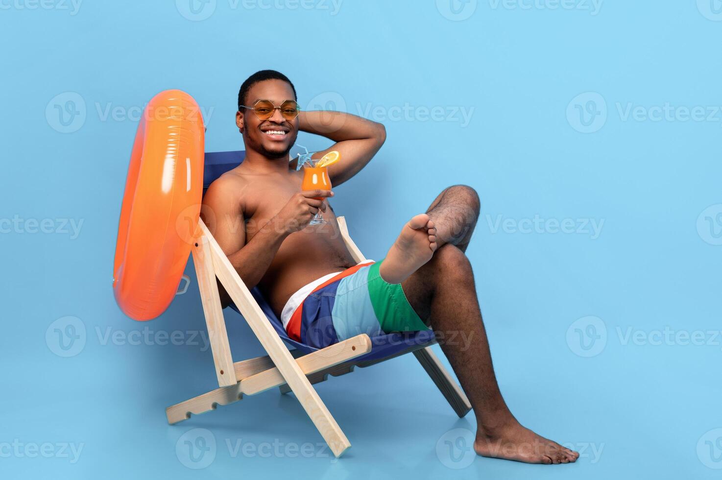 Full length of millennial black guy drinking tropical cocktail in lounge chair over blue studio background. Happy African American man chilling on beach with refreshing drink during summer vacation photo