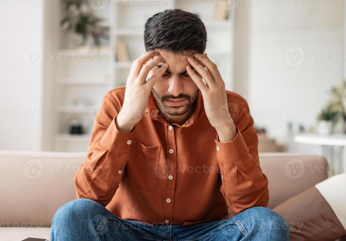 Young Middle Eastern guy suffering from headache after hard working day, sitting on couch at home, massaging head temples, having strong migraine, feeling tired, upset and stressed, copy space photo