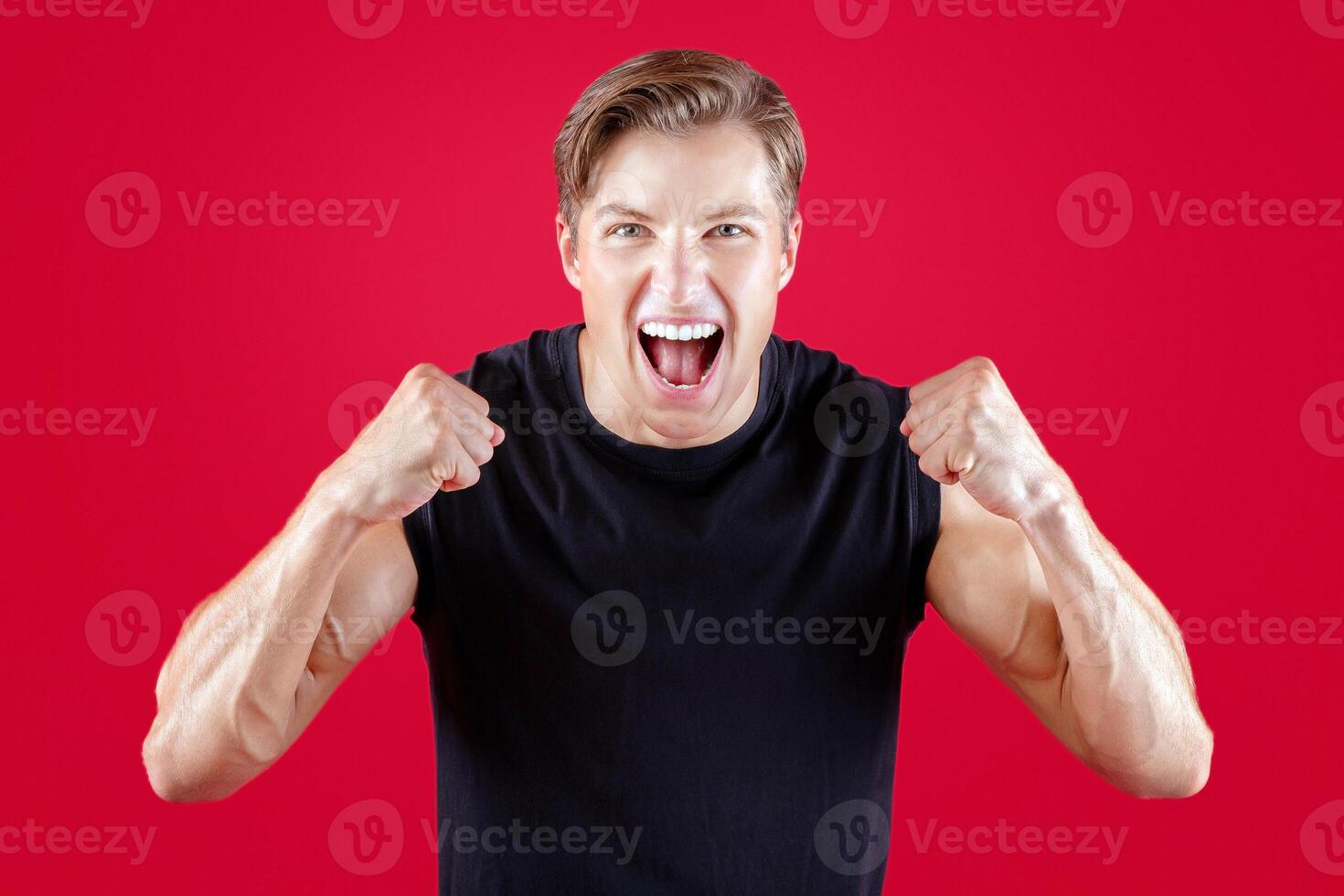 Excited millennial sportsman celebrating triumph, clenching fists, gesturing YES over red studio background. Attractive young athlete shouting in excitement, winning competition photo
