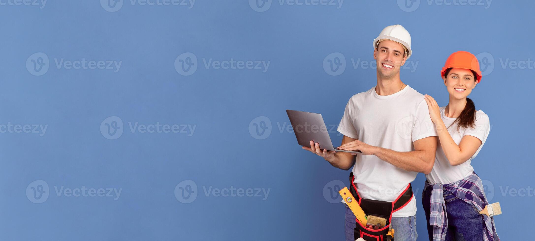 Portrait Of Happy Young Man And Woman In Hard Hats With Laptop Computer Shopping Construction Material And Repair Tools Online, Standing Together Over Blue Background, Looking At Camera, Copy Space photo