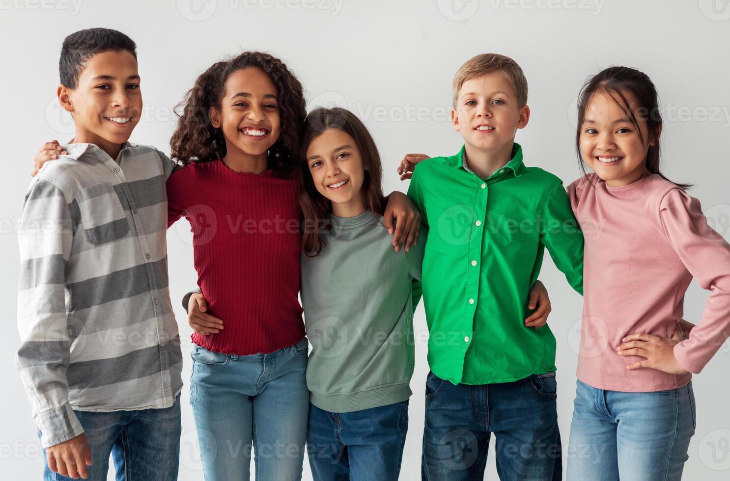 Cheerful Multicultural Preteen Kids Standing Hugging And Smiling To Camera Posing Together Over Gray Studio Background. Shot Of Mixed School Boys And Girls Group. Friendship Concept photo